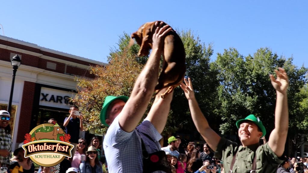 Two people in green hats lift a brown dachshund in the air during an outdoor Oktoberfest event, with a crowd watching and the "Oktoberfest Southlake" logo in the corner. Trees and a building are in the background.