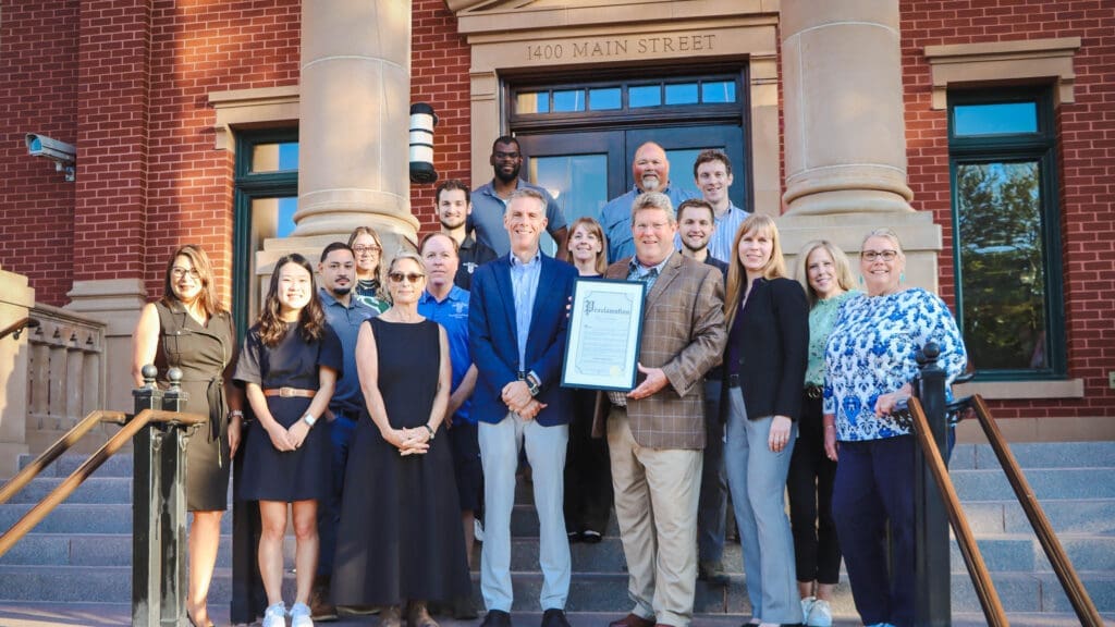 A group of people poses on the steps of a brick building, with two men in the front center holding a framed certificate. The group is smiling and standing under a sign that reads "1400 Main Street.