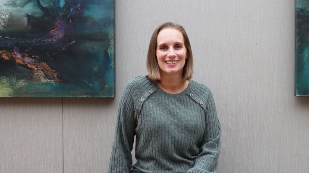 A woman with shoulder-length brown hair, wearing a grey sweater, sits and smiles in front of a light-colored textured wall with abstract paintings in the background.