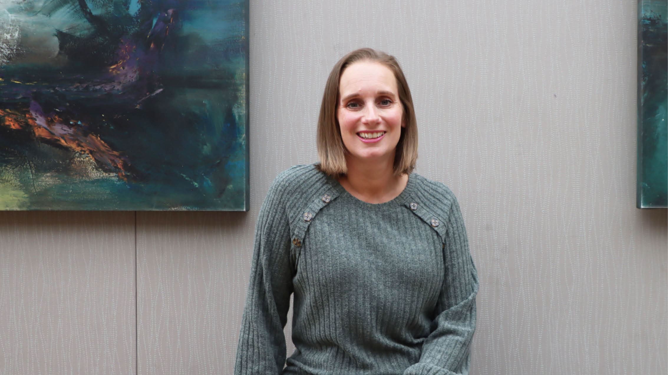 A woman with shoulder-length brown hair, wearing a grey sweater, sits and smiles in front of a light-colored textured wall with abstract paintings in the background.