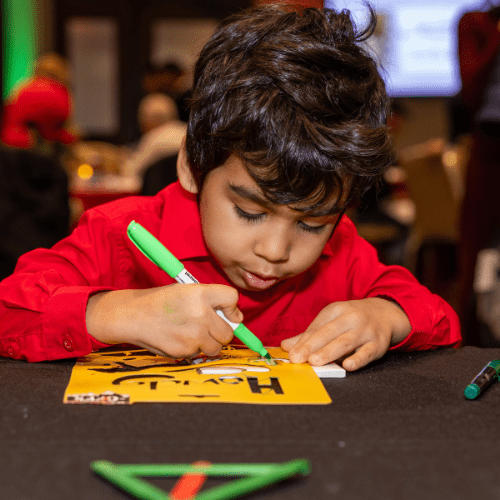 A young boy in a red shirt focuses intently as he decorates a yellow card with a green marker, sitting at a table with craft supplies.