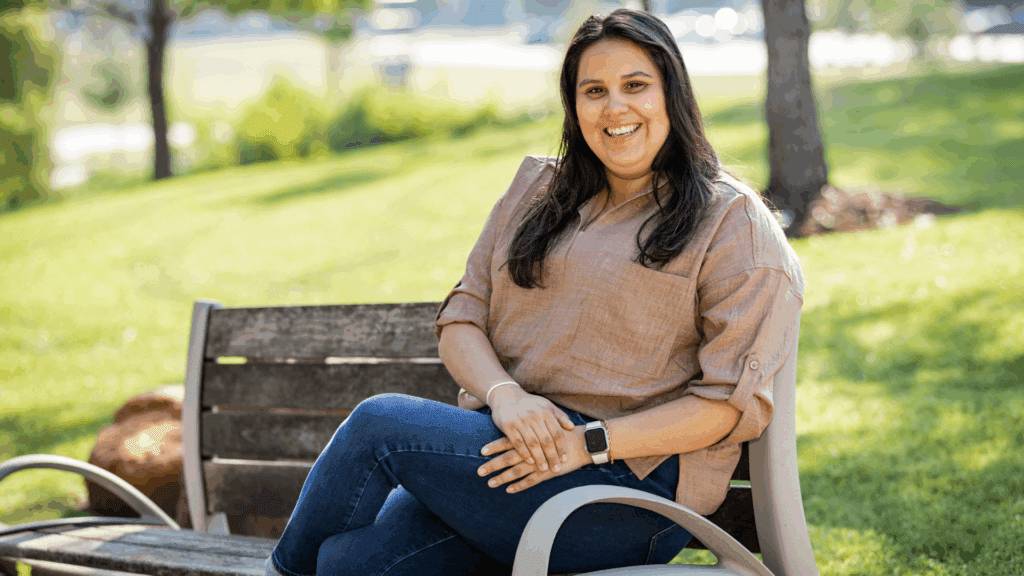 A woman with long dark hair, wearing a light brown blouse and jeans, sits smiling on a wooden park bench, surrounded by green grass and trees on a sunny day.