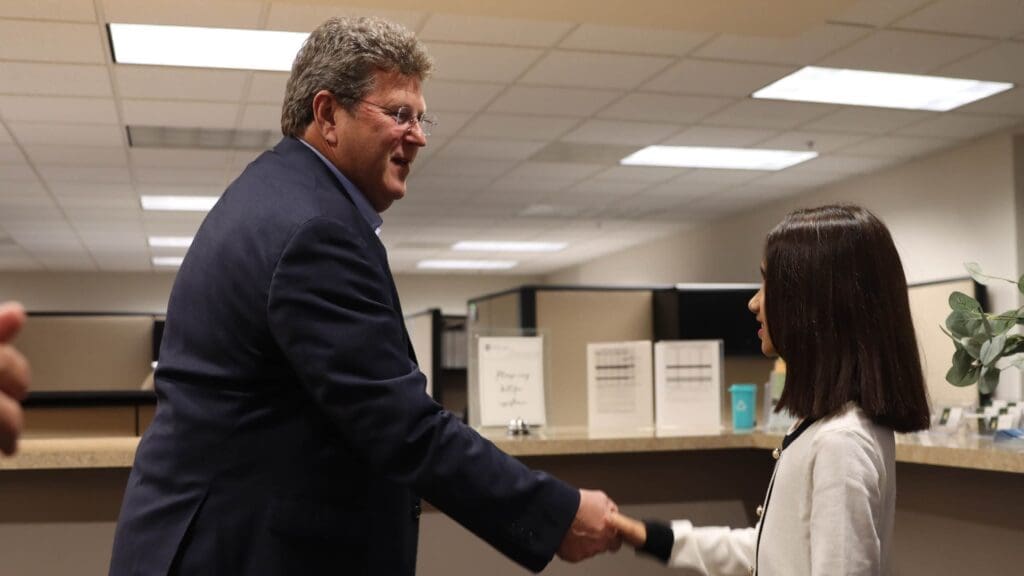A man in a suit and a woman in a white jacket are shaking hands in an office setting with cubicles and signs in the background.