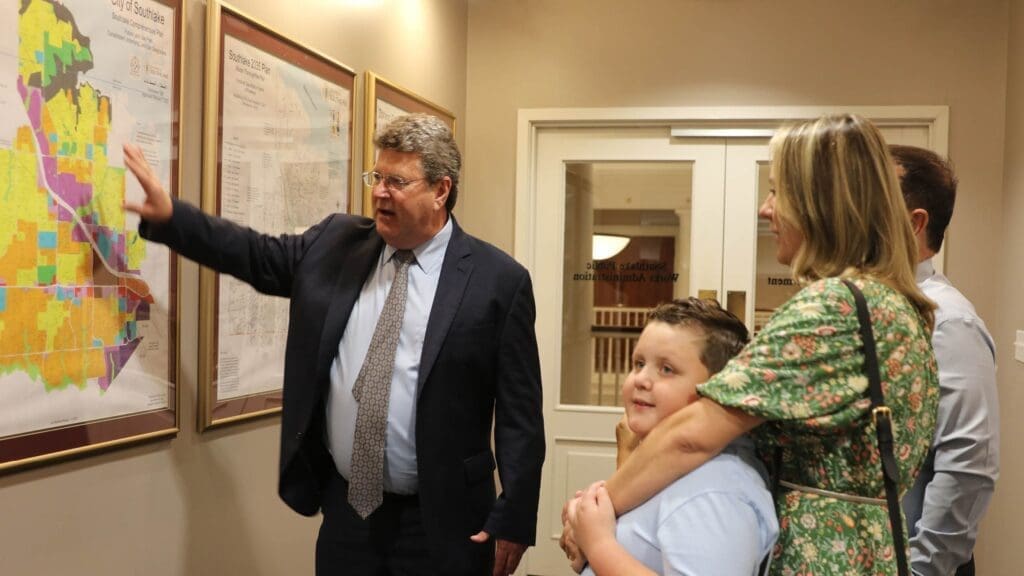 A man in a suit points to a colorful map on the wall while talking to a woman holding a boy, both looking at the map. They are standing in a well-lit hallway with framed maps and documents.
