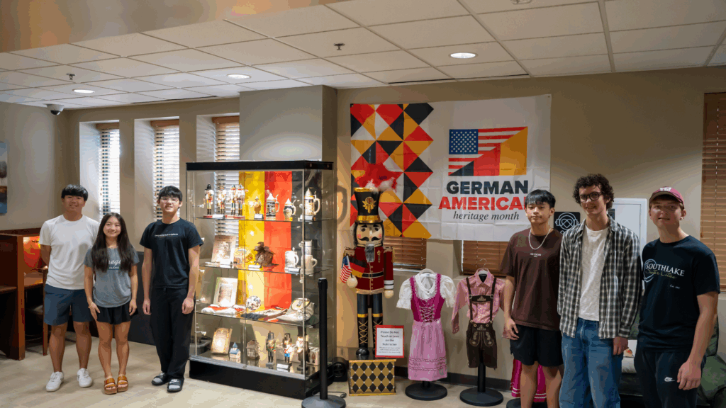 Six young people stand next to a display case of German cultural items and costumes, with a “German American Heritage Month” banner on the wall behind them in a well-lit room.