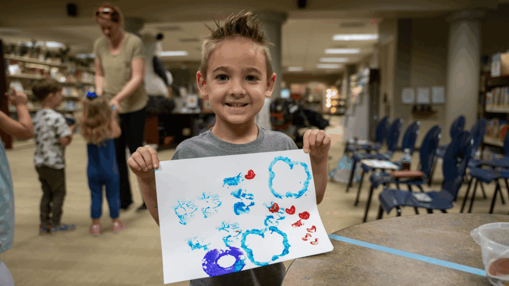 A young boy smiling and holding up a piece of paper with colorful painted shapes in a classroom or library setting. Other children and an adult are visible in the background.