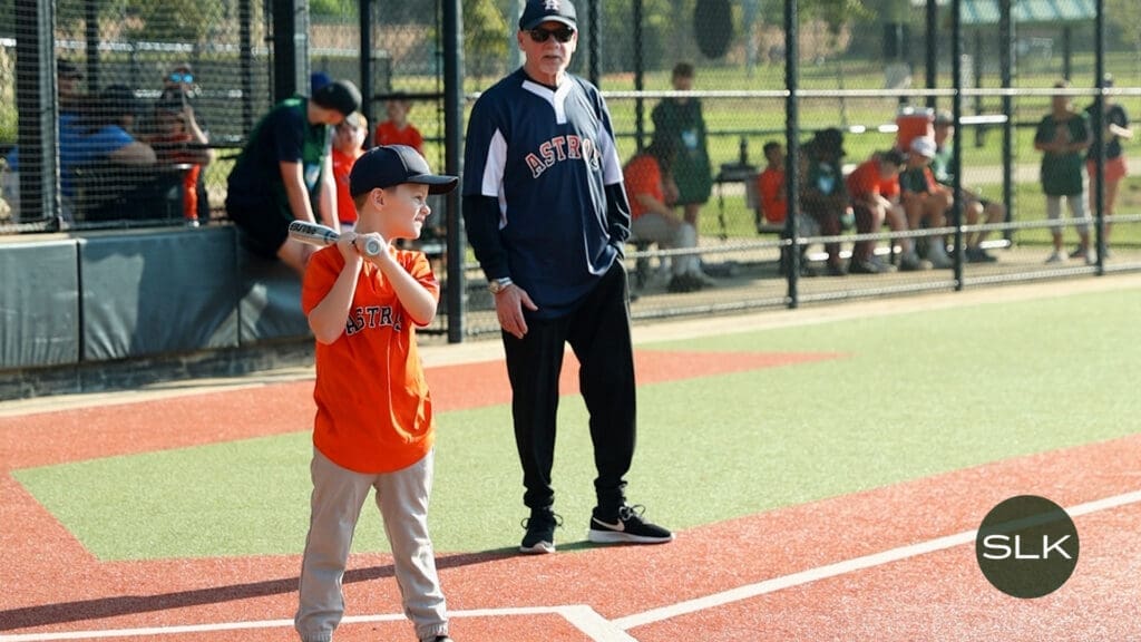 A young boy in an orange Astros shirt stands ready to bat at a baseball field, while a Coach in an Astros jersey watches nearby. Other children and adults are visible behind a fence in the background.