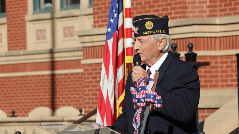 An older man in a military-style cap labeled "Southlake, TX" speaks at a podium decorated with patriotic ribbons, standing outside near a large American flag and a red brick building.