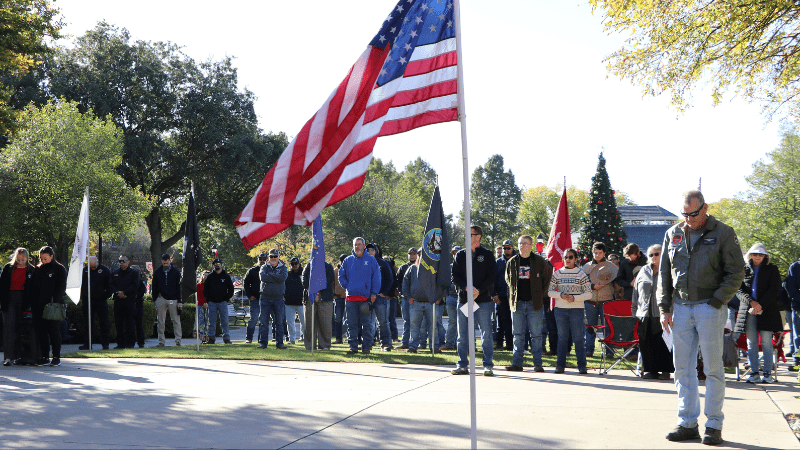 A group of people stand outdoors on a sunny day, gathered around several flagpoles, including one with an American flag in the foreground. Some individuals have their heads bowed in apparent reflection or remembrance.