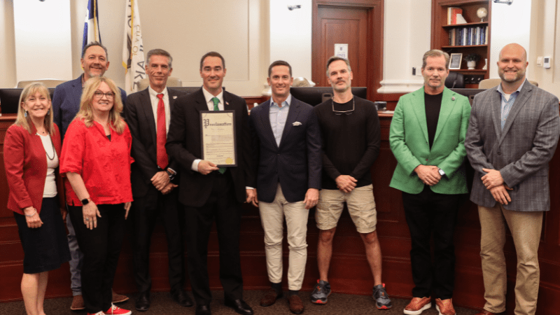 A group of nine people, dressed in business and casual attire, stand side by side in a formal room. One person in the center holds a framed document, and several people are smiling at the camera.