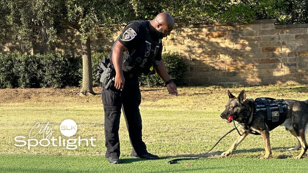 A police officer in uniform interacts with a German Shepherd wearing a harness and holding a red toy, outdoors on grass near a stone wall. The "City Spotlight" logo appears in the corner.