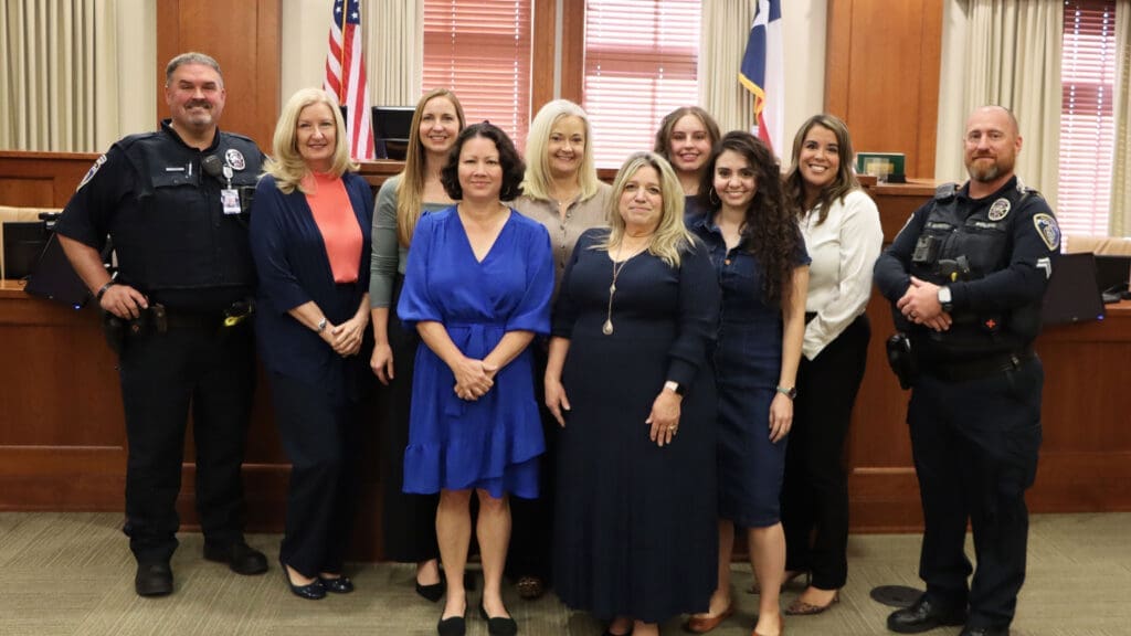 A group of ten people, including two uniformed police officers and eight women in business attire, stand together smiling in a wood-paneled room with flags and large windows in the background.