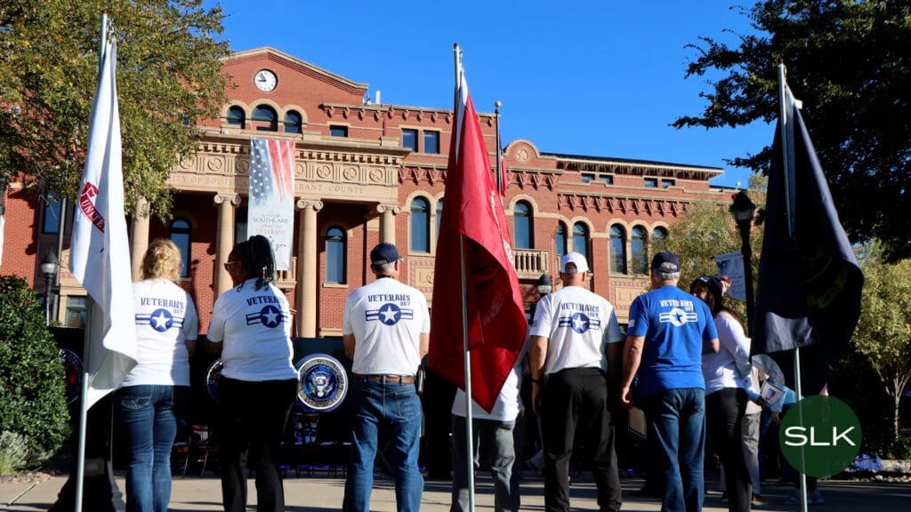 A group of people in "Veterans" shirts stand holding flags in front of Southlake Town Hall and US-themed banners, under a clear blue sky.