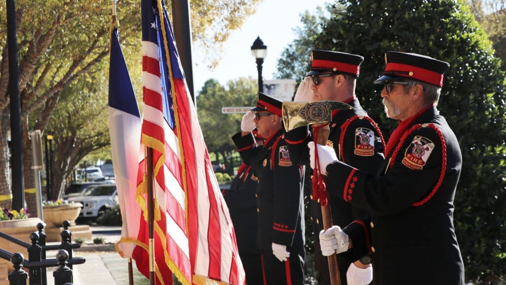 Four uniformed firefighters stand in a row during a ceremony, saluting near American and Texas flags. One firefighter holds a ceremonial axe. Trees and parked cars are visible in the background.