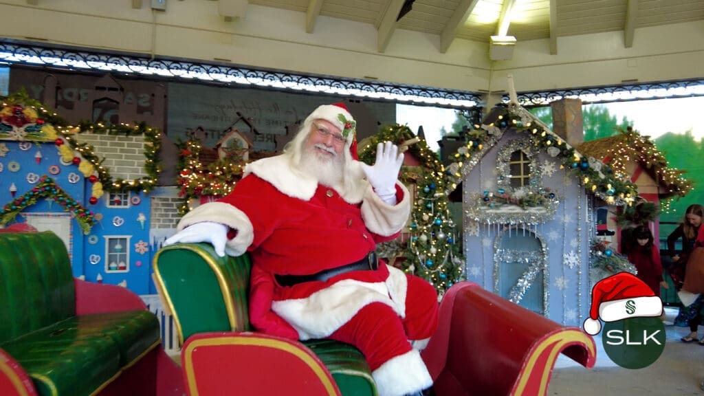 Santa Claus sits and waves from a festive sleigh, surrounded by decorated holiday houses and Christmas lights under a pavilion. The scene is cheerful and bright, capturing a joyful holiday atmosphere.