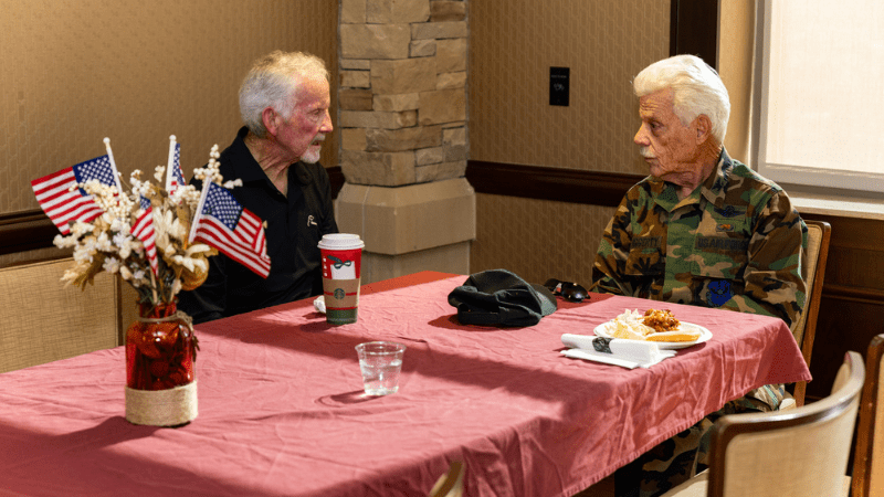 Two elderly men sit across from each other at a table with a red tablecloth. One wears a black shirt, the other wears military camouflage. American flags and flowers are in a vase on the table, along with food, a coffee cup, and a hat.