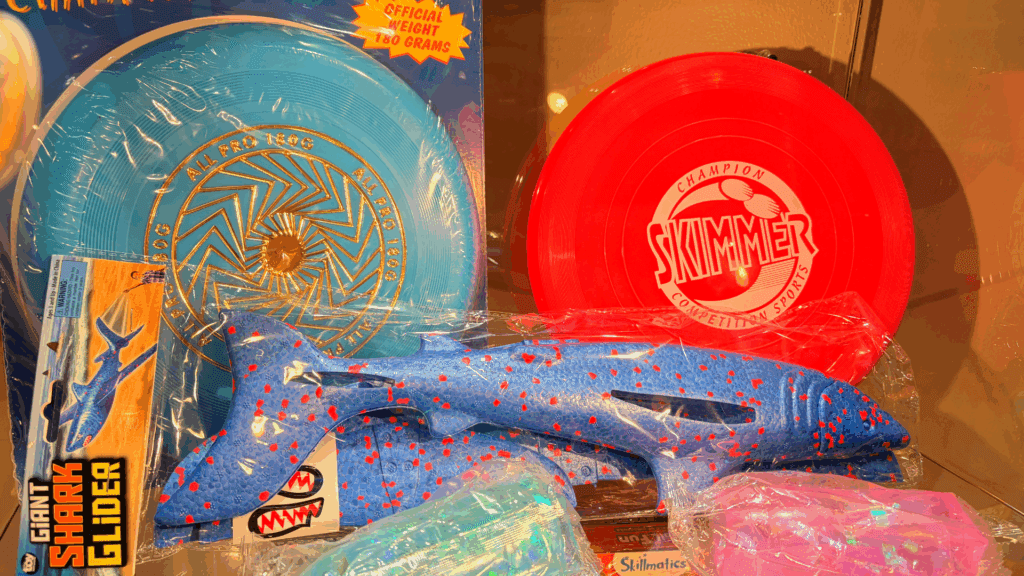 A display of colorful toys including a red and a blue flying disc, a blue toy shark with red spots, and a giant shark glider package, all wrapped in plastic. Bright lighting highlights the items.