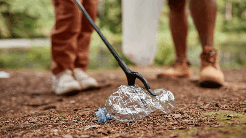 A close-up of a person using a grabber tool to pick up a crushed plastic bottle from the forest ground, with another person standing nearby, both wearing casual outdoor shoes.