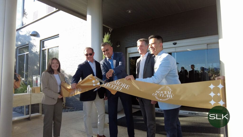Five people in business attire smile as they cut a large gold ribbon outside a building entrance during a ribbon-cutting ceremony. The ribbon has "Social Outlaw" and "More Hotel" printed on it.