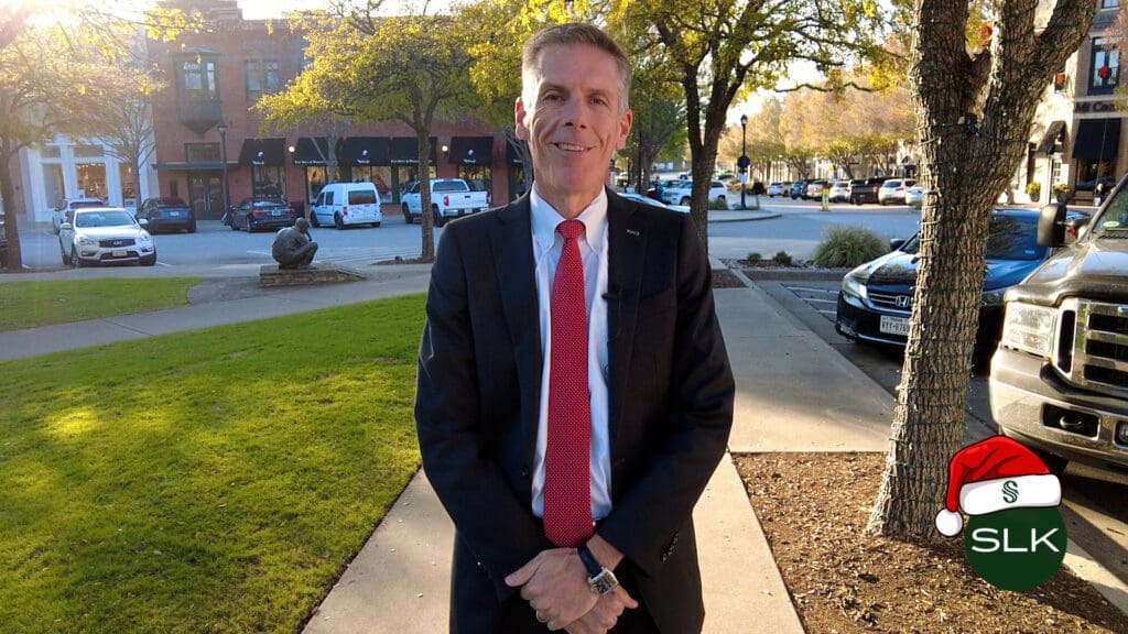 Mayor Shawn McCaskill stands in a suit and red tie on a sidewalk in Southlake Town Square area with trees and buildings in the background. The SLK logo with a Santa hat is in the lower right corner.