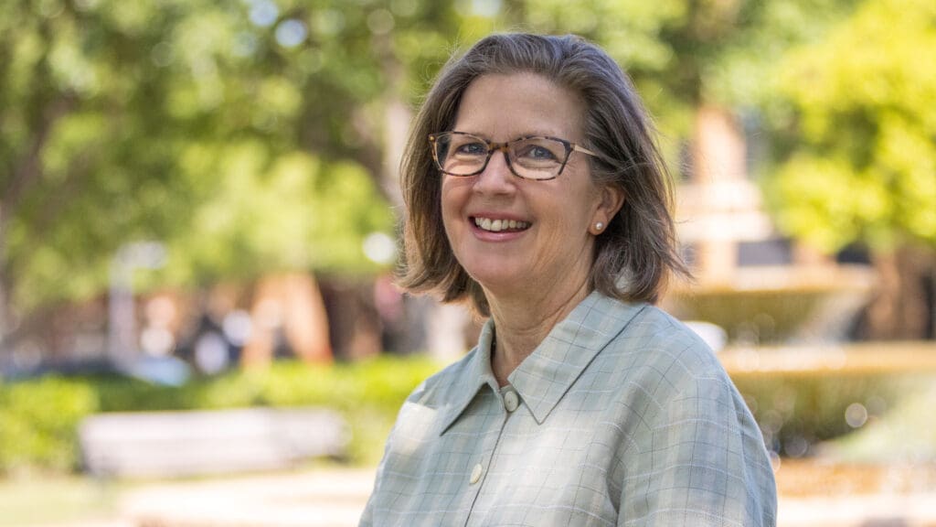 A woman with short hair and glasses smiles outdoors. She wears a light-colored, plaid button-up shirt. The background is blurred, showing trees, greenery, and a fountain.