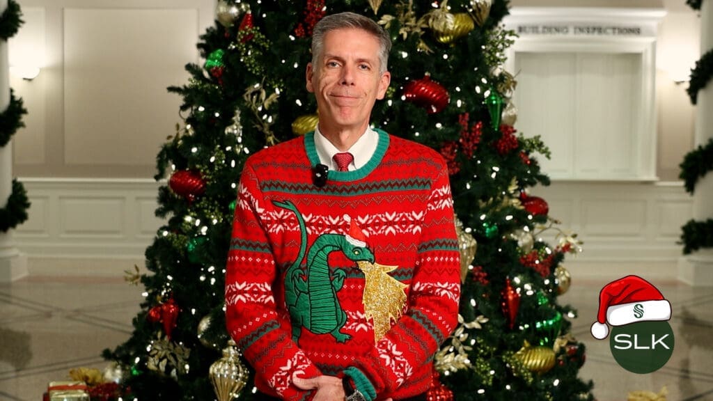 Mayor McCaskill wears a festive red holiday sweater with a green dragon stands in front of a decorated Christmas tree, smiling. The SLK logo with a Santa hat is in the lower right corner.