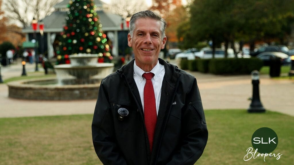 Mayor Shawn McCaskill wearing a black jacket and red tie smiles while standing in a park with a decorated Christmas tree and fountain in the background. The logo "SLK Bloopers" appears in the bottom right corner.
