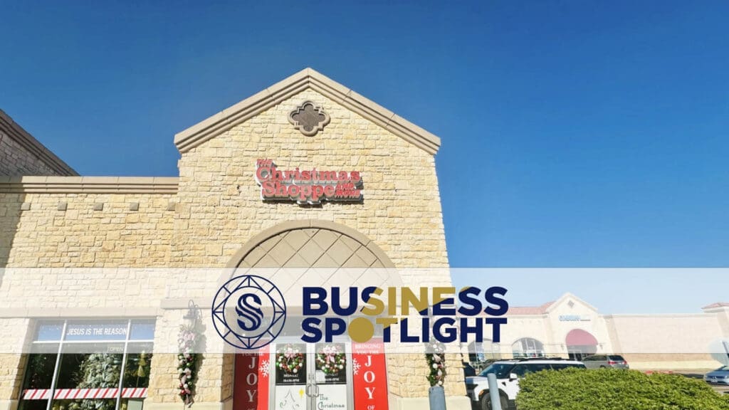 A stone shopping center with a sign reading "The Christmas Shoppe." A "Business Spotlight" banner is overlaid at the bottom. The sky is clear and blue, and cars are parked in front of the building.