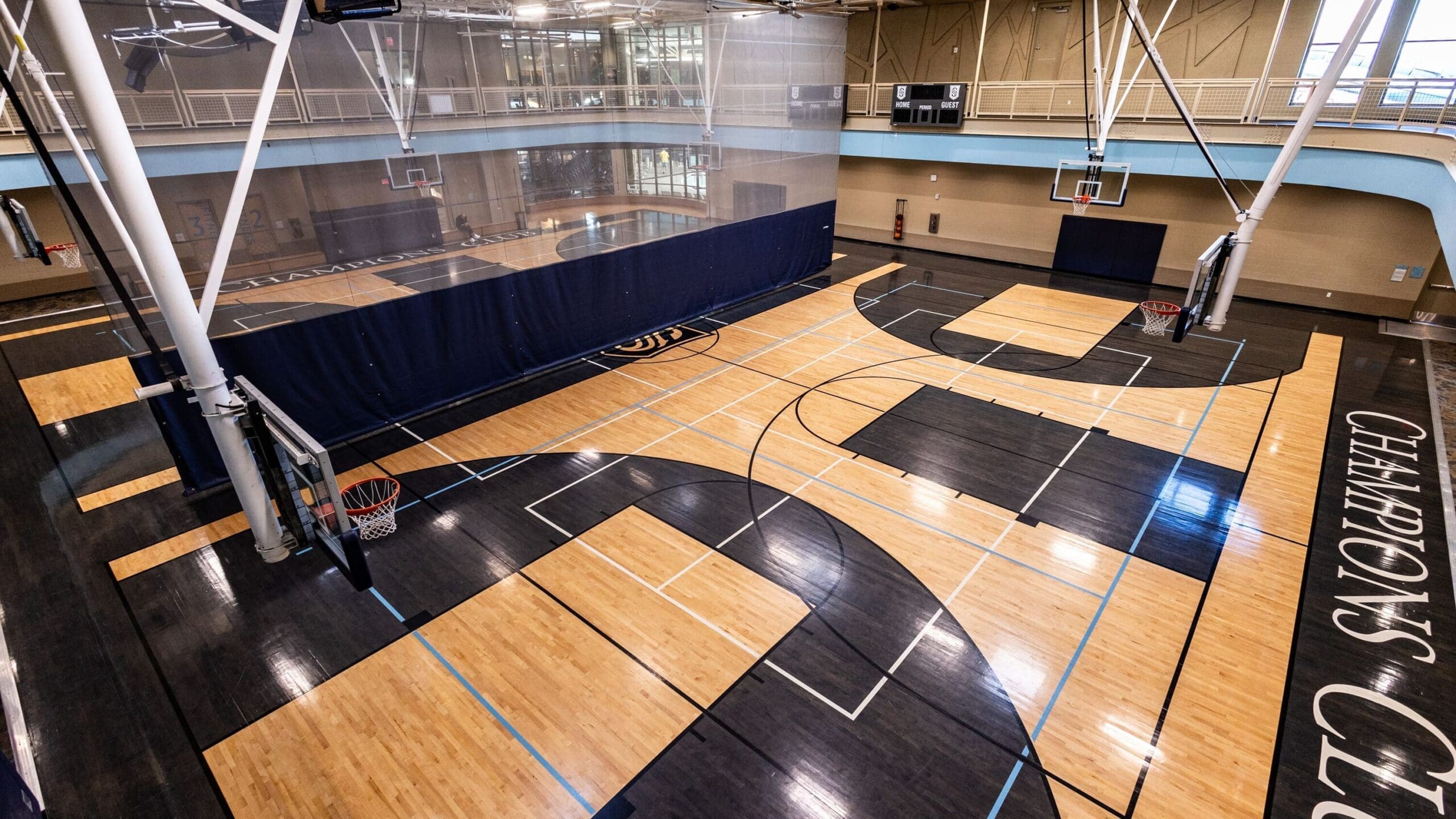 A spacious indoor gym features multiple basketball courts with wood and dark-colored flooring, blue boundary lines, six basketball hoops, and an elevated running track surrounding the courts.