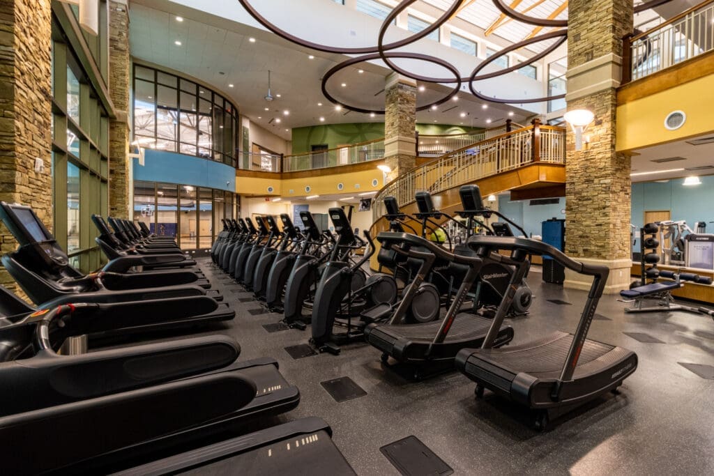 A spacious gym with rows of treadmills and elliptical machines, tall stone columns, high ceilings with modern circular lights, and a wooden staircase leading to an upper level.