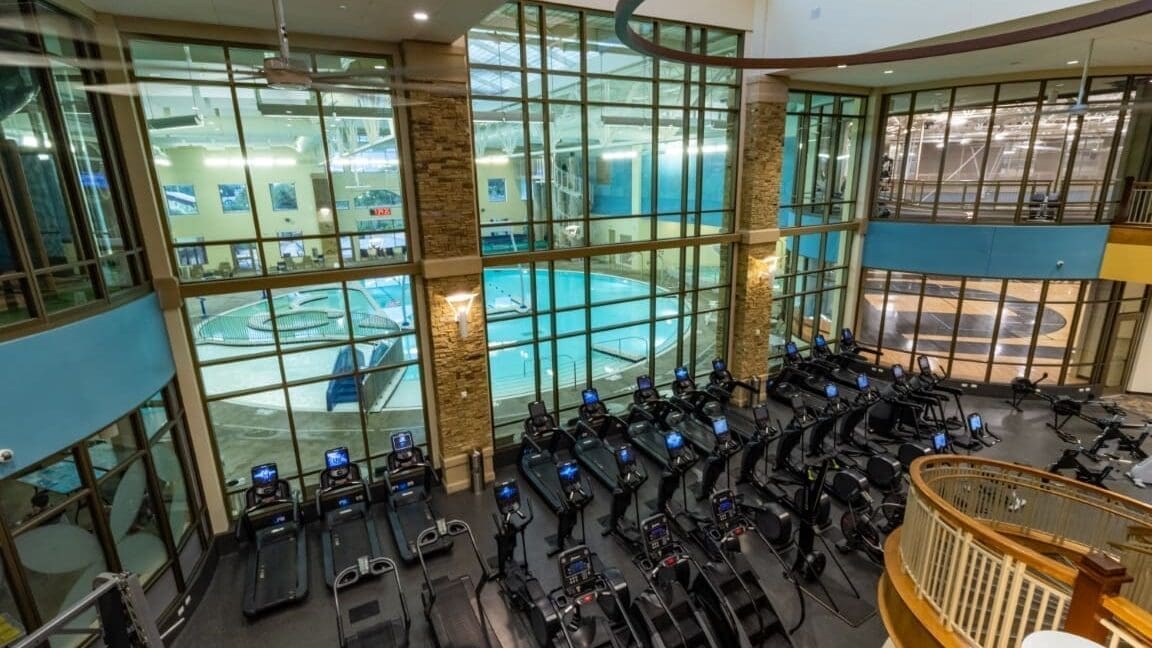 A spacious fitness center with rows of treadmills and other cardio machines faces large windows overlooking an indoor swimming pool. The area is well-lit with modern decor and glass walls.