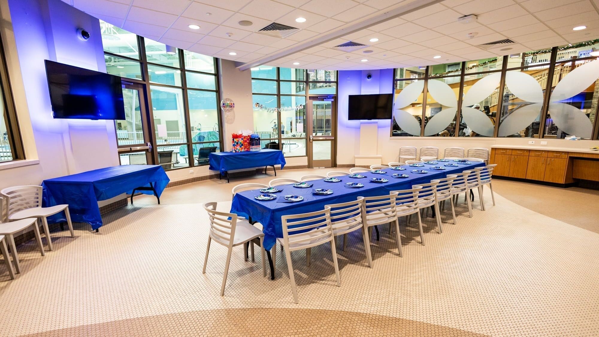 A brightly lit room with large windows, a long table covered with a blue tablecloth set for a party, surrounded by white chairs, and additional tables with blue cloths along the walls.