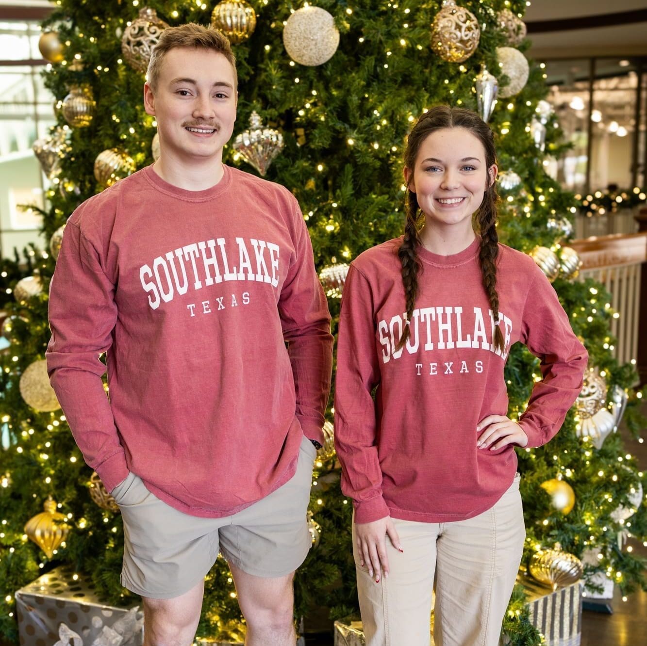 Two people stand in front of a decorated Christmas tree, both wearing matching red "Southlake Texas" long-sleeve shirts. The tree has gold ornaments and wrapped gifts underneath. They are smiling at the camera.