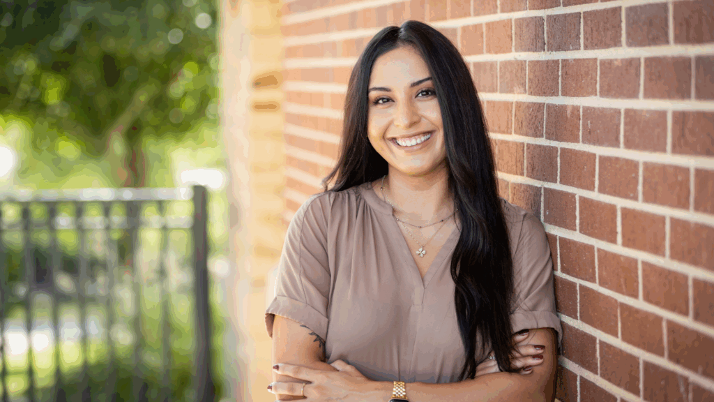 A woman with long dark hair and a light brown blouse smiles while leaning against a red brick wall outdoors, with greenery and a black fence in the background.