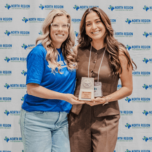 Two women smiling and holding a small award together, standing in front of a step-and-repeat banner with the "North Region" logo. One wears a blue shirt and glasses, the other a brown outfit with a name badge.