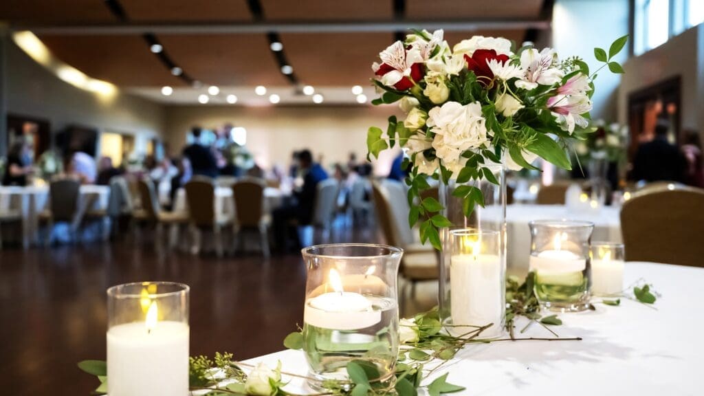 A decorated table at a formal event or wedding, featuring lit candles in glass holders, a floral arrangement with white and red flowers, and blurred guests seated at tables in the background.