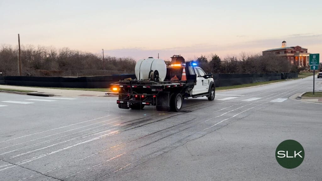 A white Southlake Public Works utility truck with flashing lights sprays brine solution on a road at dawn, leaving wet streaks. Trees, a building, and traffic signs are in the background. "SLK" watermark is in the lower right corner.
