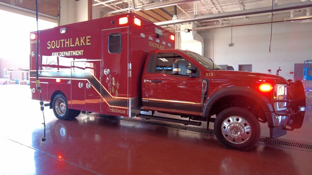 A red Southlake Fire Department ambulance is parked inside a spacious, brightly lit garage with open doors. The vehicle has emergency lights on top and reflective gold lettering on the side.