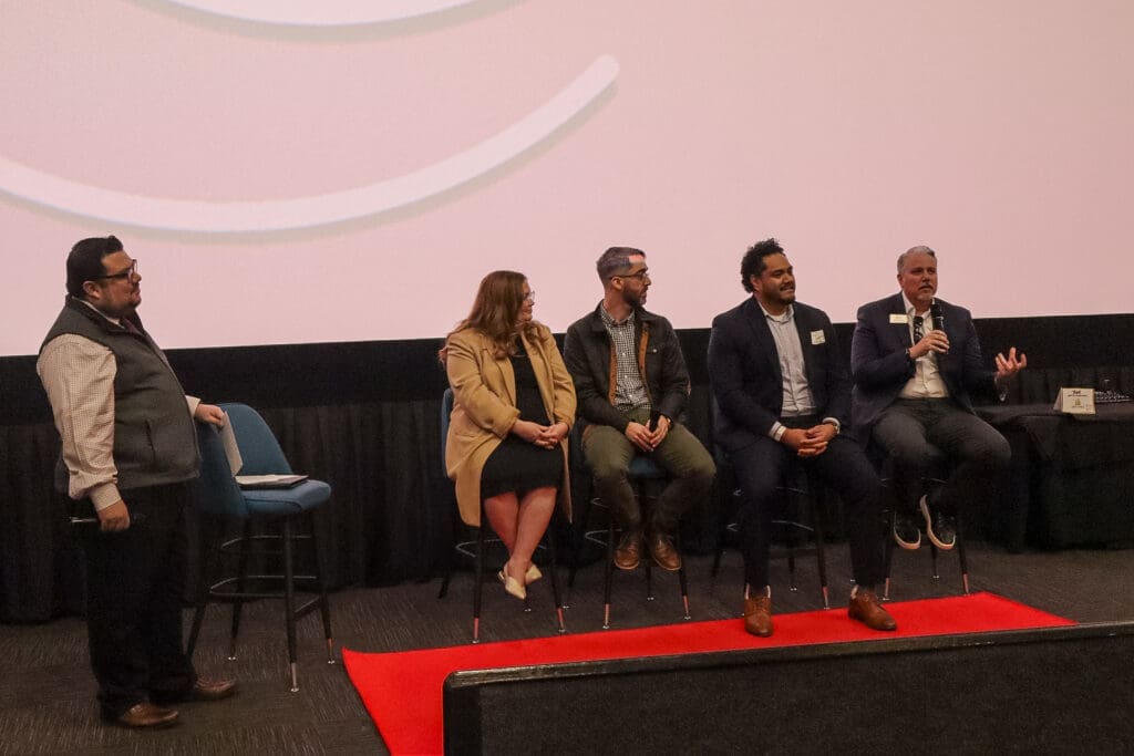 Five people participate in a panel discussion; one stands holding a microphone while four others sit on chairs, engaged and speaking, in front of a large projection screen.