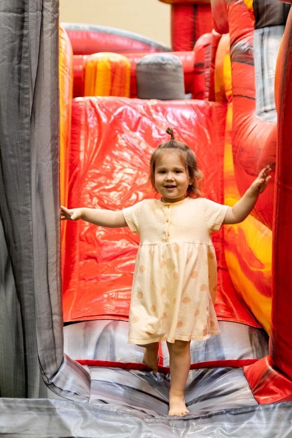 A young child in a light dress walks barefoot and smiling through an inflatable play structure with red and gray sections.