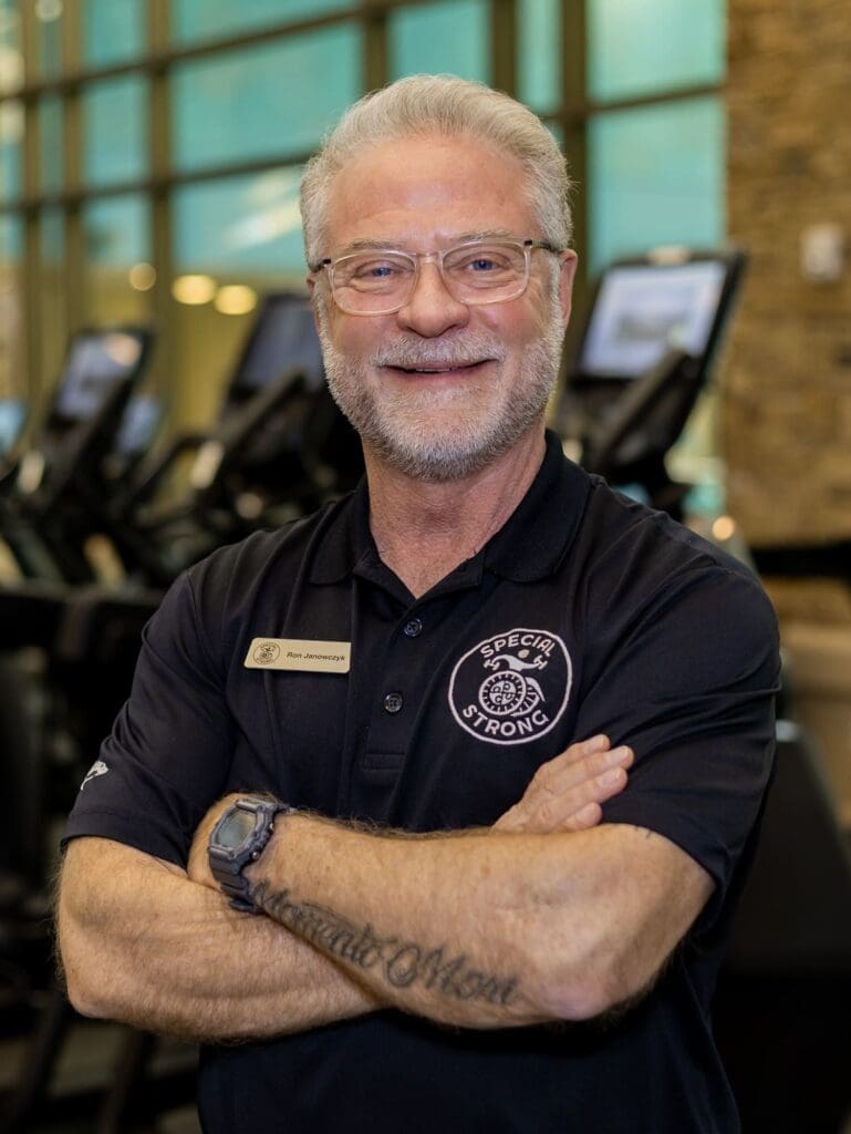 A smiling older man with gray hair and glasses stands with arms crossed in a gym, wearing a black polo shirt with a fitness logo and nametag. Cardio machines and large windows are visible in the background.