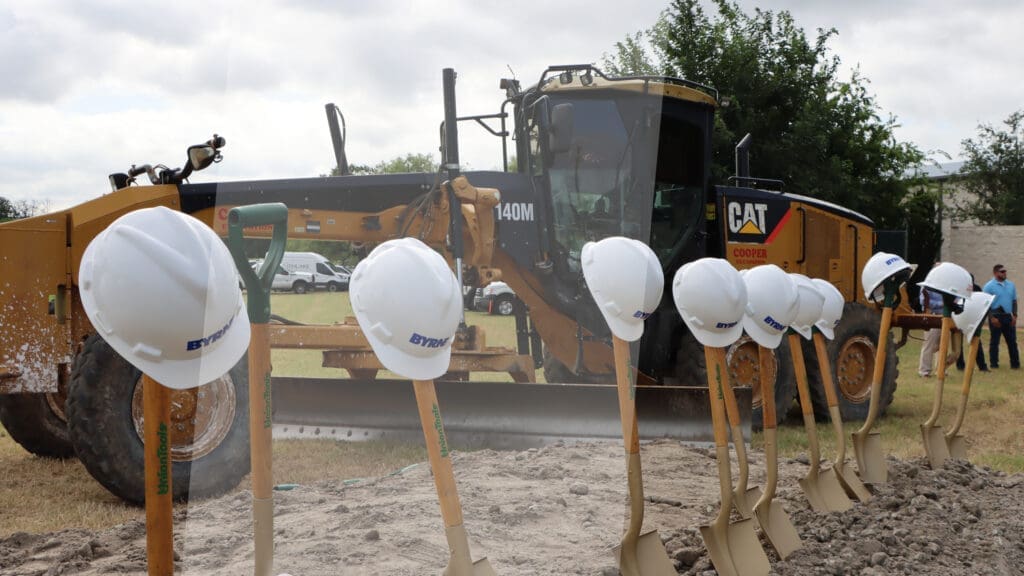 A row of shovels with white hard hats rests in the dirt at a construction site, with a large yellow Caterpillar grader machine in the background and several people standing nearby.