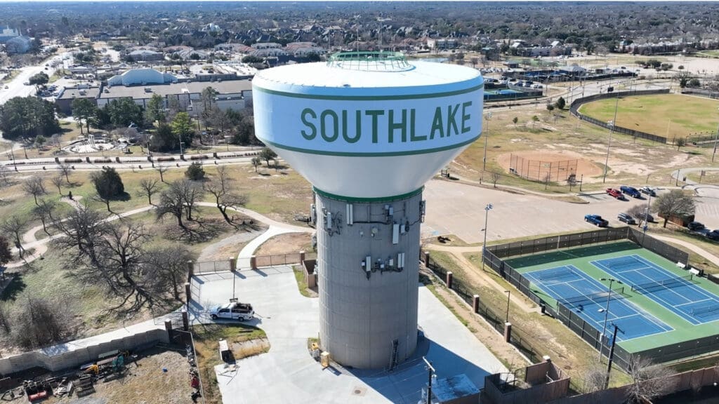 An elevated water tower labeled "SOUTHLAKE" stands in a suburban area, surrounded by trees, tennis courts, parking lots, and nearby buildings on a clear day.