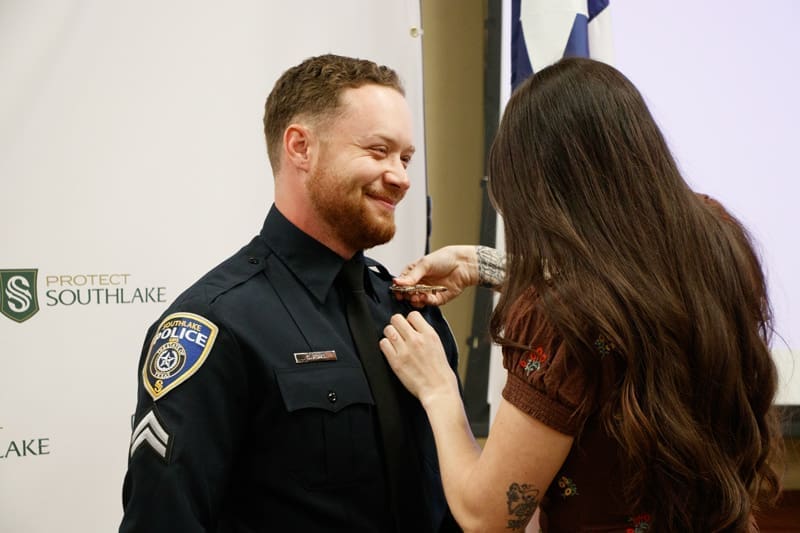 A smiling police officer in uniform is having a pin attached to his collar by a woman with long hair during a ceremony. A "Protect Southlake" banner is visible in the background.