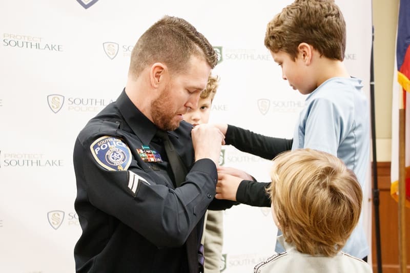 A police officer kneels and interacts with three young boys, one of whom is pinning a badge on the officer's uniform. The background features police and city logos.