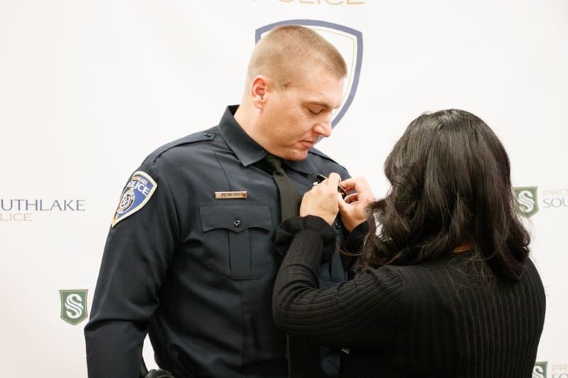 A woman pins a badge onto the uniform of a male police officer during a ceremony. The officer stands facing her, and both appear focused on the task. The background shows police and city logos.