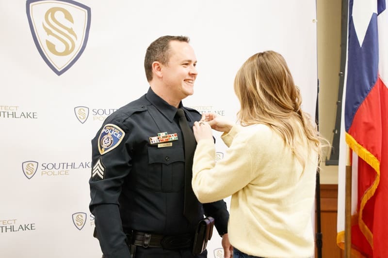 A woman pins a badge onto a smiling police officer’s uniform during a ceremony. They stand in front of a Southlake Police banner and beside a Texas state flag.