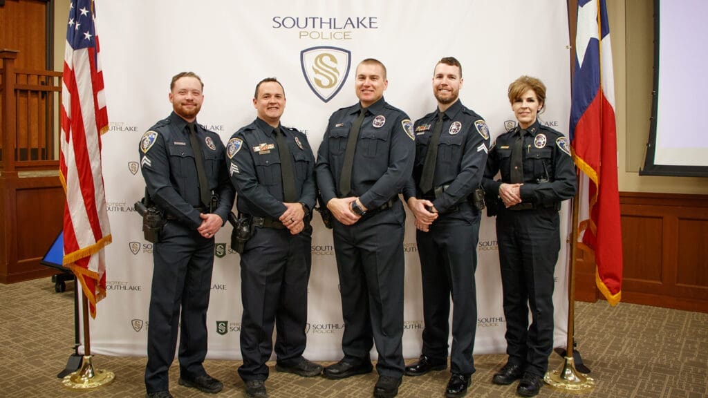 Five uniformed police officers stand in front of a "Southlake Police" backdrop, posing for a group photo. Two flags are visible on the sides and the group is indoors on a carpeted floor.
