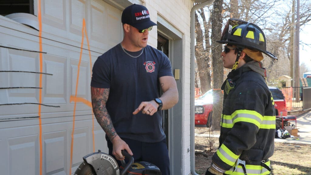 Two firefighters stand outside a garage. One, in a navy shirt, is holding a saw and giving instructions to the other, who is wearing full firefighting gear. Orange markings are visible on the garage door.