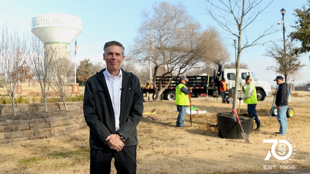 Mayor Shawn McCaskill smiling in a park while workers in safety vests plant trees in the background. A water tower labeled "Southlake" and a crew planting a tree are seen in the background. The image has a "70 Years Est. 1956" logo in the bottom right corner.